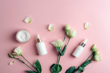 White cosmetic serum bottles, cream jar and eustoma flowers on pink background. Top view, flat lay.