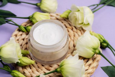 Cosmetic cream jar and white eustoma flowers on a wicker coaster on purple background. Top view.