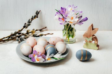 Boyanmış Paskalya yumurtaları ve tahta oyuncak tavşancığı olan gri tabak, beyaz masadaki krokus çiçekleri. Paskalya tebrik kartı.