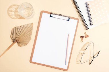 Boş pano, not defteri, yelpaze yaprağı, gözlük ve güneş ışığında açık bej arkaplanda bir bardak su. Üst görünüm, düz görünüm, model.