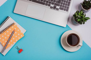 Bir fincan kahve, dizüstü bilgisayar, not defterleri, kalem ve sulu bitkiler mavi arka planda. Rahat bir iş yeri, ev konsepti işi. Üst görünüm, düz görünüm, kopyalama alanı.