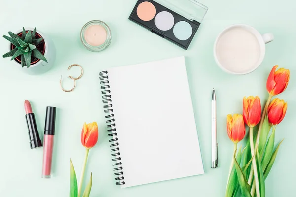 stock image Blank notepad, cup of coffee, make up cosmetics and bouquet of orange tulips on light green background, top view. Womens day or Mothers day concept. Creative flat lay, mockup.