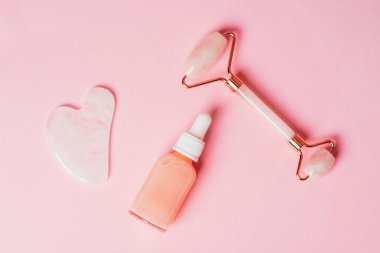Gül kuvars kristal yüz silindiri ve gua sha kazıyıcı, yüz serumu veya pembe arka planda organik kozmetik yağ. Kaldırma terapisi için yüz masajı kiti. Cilt bakımı yaşlanmayı önleyici aletler. Üst manzara, düz uzanma.