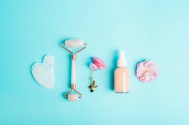 Serum şişesi, yüz silindiri, gua sha taşı ve mavi arka planda pembe sakura çiçeği. Cilt bakımı, güzellik tedavi konsepti. Üst manzara, düz uzanma.