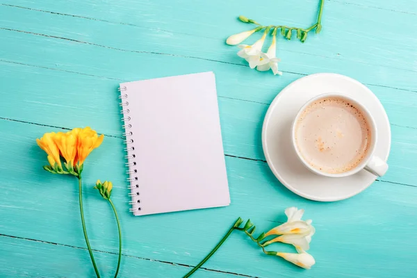 Blank notepad and coffee cup on turquoise wooden table with freesia flowers. Top view, flat lay, mockup.