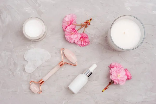 stock image Cream jar, face roller, gua sha stone and pink cherry blossom on gray concrete background. Skin care, beauty treatment concept. Top view, flat lay.