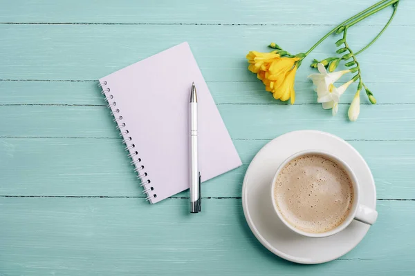 stock image Blank notepad, pen and coffee cup on turquoise wooden table with freesia flowers. Top view, flat lay, mockup.