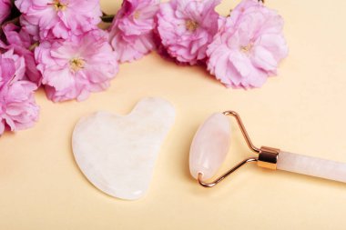 Yüz silindiri, gua sha taşı ve bej arka planda pembe kiraz çiçeği. Cilt bakımı, güzellik tedavi konsepti. Üst görünüm.