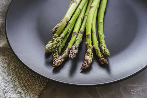 stock image Fresh green asparagus on black plate on grey concrete table. Healthy vegan raw food concept. Closeup.