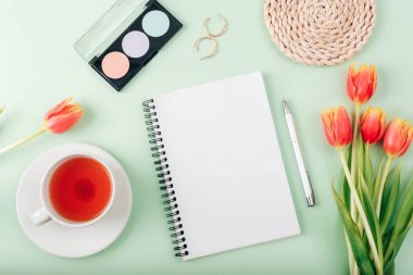 Boş not defteri, bir fincan çay, makyaj malzemesi ve açık yeşil arka planda bir buket kırmızı lale, üst manzara. Kadınlar Günü ya da Anneler Günü konsepti. Yaratıcı düz döşeme, model.