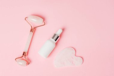 Gül kuvars kristal yüz silindiri ve gua sha kazıyıcı, yüz serumu veya pembe arka planda organik kozmetik yağ. Kaldırma terapisi için yüz masajı kiti. Cilt bakımı. Üst görünüm, düz görünüm, kopyalama alanı.