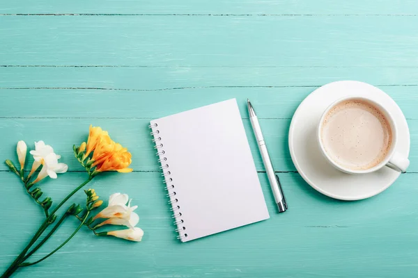 stock image Blank notepad, pen and coffee cup on turquoise wooden table with freesia flowers. Top view, flat lay, mockup.