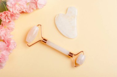 Yüz silindiri, gua sha taşı ve bej arka planda pembe kiraz çiçeği. Cilt bakımı, güzellik tedavi konsepti. Üst manzara, düz uzanma.
