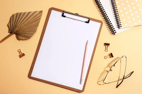 Desktop with blank clipboard, notepads, fan leaf, eyeglasses and accessories on neutral beige background. Top view, flat lay, mockup.