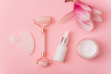 Gül kuvars kristal yüz silindiri ve gua sha kazıyıcı, yüz serumu veya organik kozmetik yağ, pembe arka planda manolya çiçeği. Kaldırma terapisi için yüz masajı kiti. Üst manzara, düz uzanma.