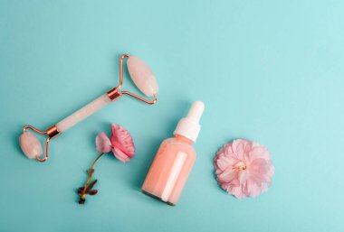 Serum şişesi, yüz silindiri ve mavi arka planda pembe sakura çiçeği. Cilt bakımı, güzellik tedavi konsepti. Üst görünüm, düz görünüm, kopyalama alanı.