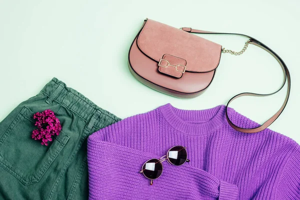 Stylish woman's outfit: purple sweater, green jeans, sunglasses and handbag. Lilac flowers decoration. Top view, flat lay.