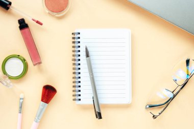 Kalem, gözlük, defter ve makyaj malzemeleriyle bej arka planda boş not defteri. Güzellik blogcusu kavramı. Üst manzara, düz uzanma.