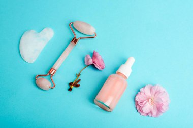 Serum şişesi, yüz silindiri, gua sha taşı ve turkuaz mavi arka planda pembe sakura çiçeği. Cilt bakımı, güzellik tedavi konsepti. Üst görünüm.