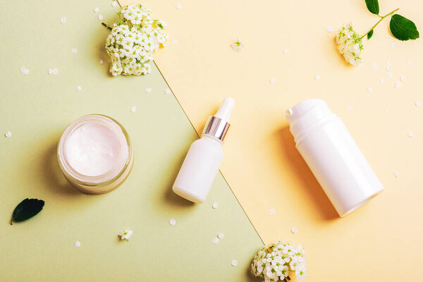 Cosmetic cream jar, serum bottle and dispenser with face cleanser, small white flowers on green and yellow background. Skin care concept. Top view, flat lay, mockup.
