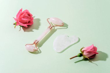 Yüz silindiri, gua sha taşı ve açık yeşil arka planda pembe gül çiçekleri ve keskin gölgeler. Cilt bakımı, güzellik tedavi konsepti. Üst manzara, düz uzanma.