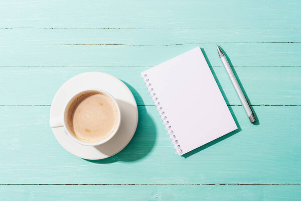 A cup of coffee, notepad and pen on turquoise blue table in sunlight. Top view, flat lay, mockup.