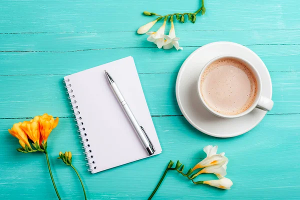 Stock image Blank notepad, pen and coffee cup on turquoise wooden table with freesia white and yellow flowers. Top view, flat lay, mockup.