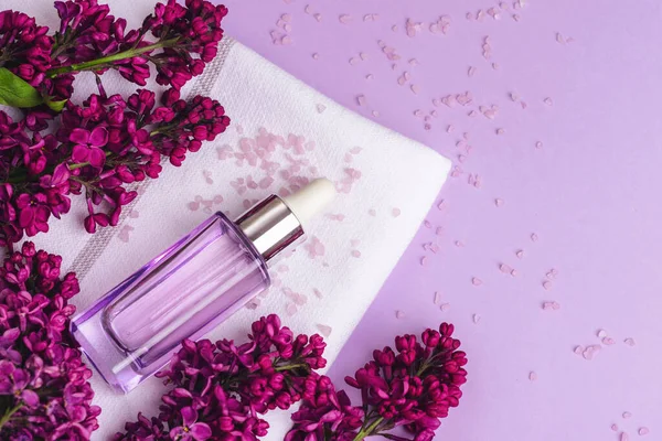 Serum bottle and lilac flowers on light purple background. Natural cosmetics, skin care concept. Top view, flat lay.