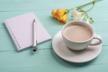 Boş not defteri, turkuaz ahşap masa üzerinde beyaz ve sarı çiçekli kalem ve kahve fincanı. Yakın çekim..