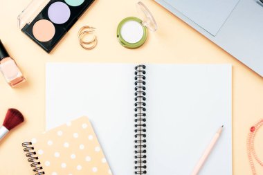 Boş not defteri, makyaj malzemesi ve pastel turuncu arka planda dizüstü bilgisayar, üst görünüm. Yaratıcı düz döşeme, model.