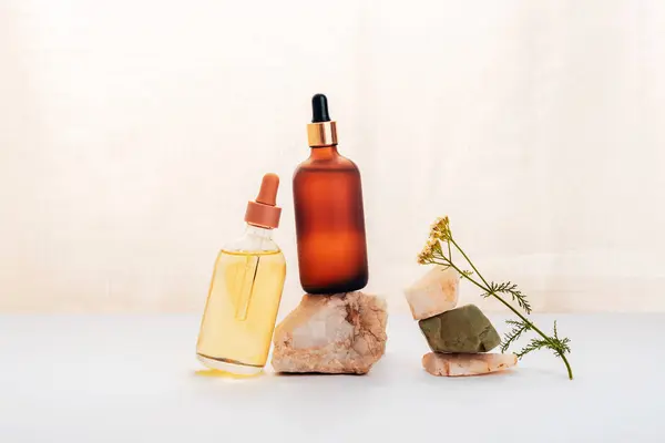 Two cosmetic oil or serum bottles on a stone and yarrow flower on white table. Natural cosmetics, spa and wellness concept. Front view.