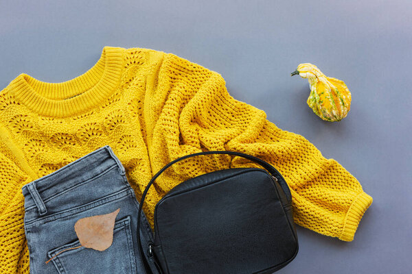 Плоский узор женского желтого свитера, серые джинсы, черная сумка и маленькая тыква, осенний лист на сером фоне. Осенняя мода. Вид сверху, плоский.