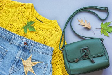 Düz kadın yatağı sarı süveter, kot pantolon, yeşil çanta ve gri arka planda sonbahar yaprakları. Sonbahar moda konsepti. Üst manzara, düz uzanma.