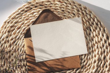 Envelope with blank card on a wicker place mat with flowers shadow in sunlight. Top view, flat lay, mockup. clipart