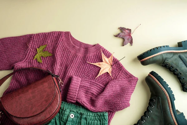Flat lay of womans purple sweater, green jeans, bag, boots and autumn leaves on neutral background. Autumn fashion concept. Top view, flat lay.
