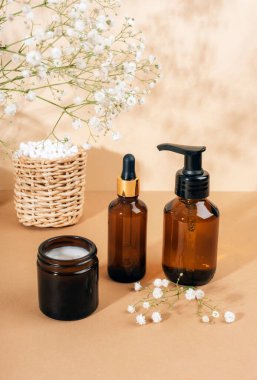 Kozmetik yüz temizleyici, serum kahverengi cam şişeler, krema kavanozu ve açık bej arka planda çingene beyaz çiçekleri. Doğal kozmetik, spa ve sağlık konsepti. Yakın çekim..