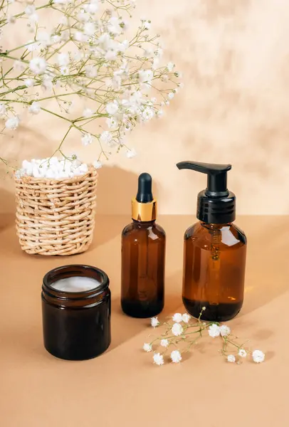 Cosmetic face cleanser, serum brown glass bottles, cream jar and gypsophila white flowers on light beige background. Natural cosmetics, spa and wellness concept. Closeup.