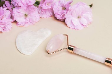 Yüz silindiri, gua sha taşı ve tarafsız arka planda pembe kiraz çiçeği. Cilt bakımı, güzellik tedavi konsepti.