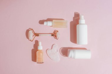 Kozmetik şişeleri ve yüz silindiri pembe arka planda gua sha taşı gün ışığında. Cilt bakımı, spa ve sağlık konsepti. Üst manzara, düz uzanma.