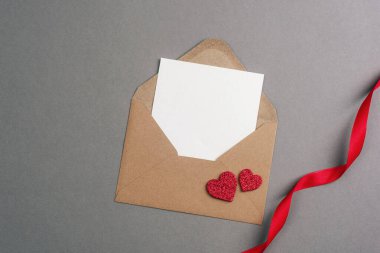 İçinde boş kart, kırmızı kalpler ve gri arka planda kurdele olan bir zarf. Sevgililer Günü konsepti, üst görünüm, düz görünüm, model.