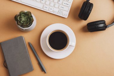 Kahve fincanı, not defteri, klavye, sulu bitki ve bej masasında kulaklık olan bir masa. Üst manzara, düz uzanma.