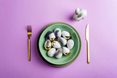 Yeşil tabaklı masa takımı, mor arka planda Paskalya yumurtası ve tavşan. Üst görünüm, düz görünüm, kopyalama alanı.