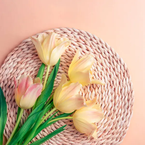 Yellow-pink tulip flowers on wicker place mat on peachy background. Spring holiday concept. Top view, flat lay.