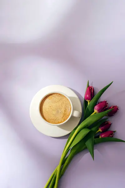 A cup of coffee and purple tulips on lilac background in sunlight. Top view, flat lay.
