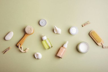 Kozmetik yağ ya da serum şişeleri, ahşap yüz fırçası, krem kavanozu ve zeytin yeşili arka planda deniz kabukları. Doğal kozmetik malzemelerle kaplıca konsepti. Üst manzara, düz uzanma.