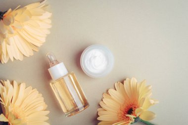 Kozmetik serumu ve krema kavanozu, gri arka planda sarı gerbera çiçekleri. Üst görünüm, düz görünüm, kopyalama alanı.