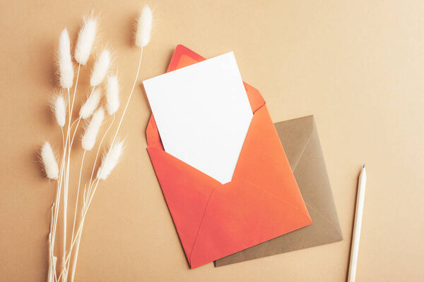 Red envelope with blank card, pencil and bunny tails grass Lagurus Ovatus flowers on brown background. Top view, flat lay, mockup.