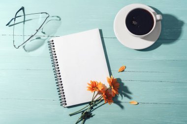 Boş not defteri, kahve fincanı ve turkuaz mavi ahşap masa üzerinde güneş ışığı altında sarı çiçekler. Üst görünüm, düz görünüm, model.