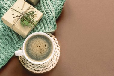 Düz bir fincan kahve, paketlenmiş hediyeler ve fotokopi alanı olan sıcak kahverengi arka planda yeşil örülmüş bir eşarp. Yıl Rengi: 2025 Mocha Mousse.
