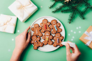 Noel kurabiyelerini sarılı beyaz bir tabakta süsleyen bir kızın elleri ve yeşil arka plandaki neşeli unsurlar. Üst manzara, düz uzanma.